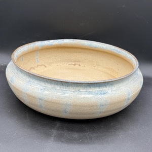 May include: A handmade ceramic bowl with a wide, shallow design. The bowl features a light beige interior and a light blue and beige exterior. The rim has a subtle blue glaze. The bowl is suitable for serving or display.