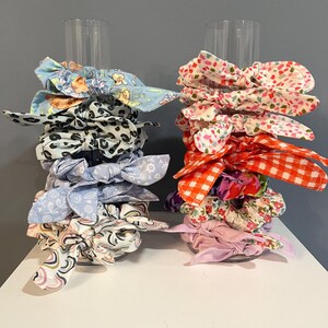 May include: Two stacks of colorful fabric scrunchies with different patterns. The scrunchies are stacked on clear acrylic stands.