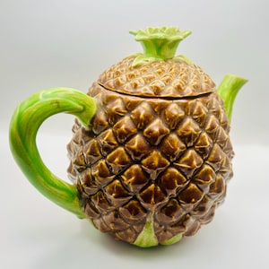 May include: A ceramic teapot in the shape of a pineapple. The teapot is brown with a textured surface, green handle, spout, and top. The lid is also brown and has a green pineapple top. The teapot is set against a white background.