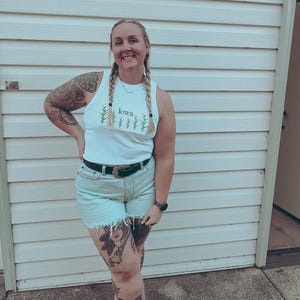 May include: A white sleeveless tank top featuring a green graphic of corn stalks and the text Iowa printed in the center. The top is paired with light-wash denim shorts with a frayed hem and a black leather belt with a silver-toned metal buckle.