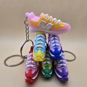 May include: A stack of colourful miniature sneaker keychains. The top keychain is yellow, pink, and white. The other keychains are red, green, purple, blue, and light pink.