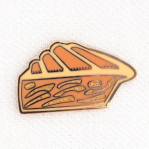 Könnte beinhalten: Eine goldfarbene Emaille-Anstecknadel in Form eines Stückes Kuchen mit einer braunen Kruste und einer gelben Füllung. Die Füllung hat eine glitzernde Textur.