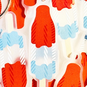 May include: A selection of popsicle-shaped keyrings. Each keyring has a red, white, and blue design with a popsicle stick. The keyrings are made of clear acrylic and have a gold-coloured keyring.