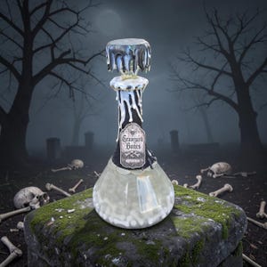 May include: A clear glass decanter filled with small white bone-shaped objects. The neck is wrapped in black fabric with white wax-like drips extending from the stopper. A label on the front reads Graveyard Bones in black gothic lettering.