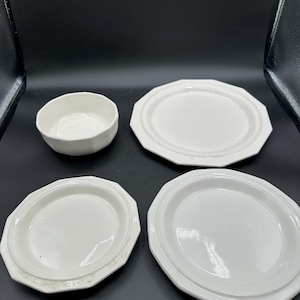 May include: A set of four white ceramic dishes consisting of one small bowl and three plates. Each piece features a smooth, glossy finish and a distinct octagonal rim design. The items are arranged in a group, showcasing their matching off-white color and uniform, clean-lined geometric shape throughout the collection.