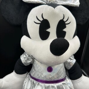K&ouml;nnte beinhalten: Ein Pl&uuml;sch Minnie Mouse-Puppe mit silberner Schleife und Kleid mit wei&szlig;en Punkten. Die Puppe hat schwarze Ohren, Augen und Nase sowie wei&szlig;e Handschuhe. Ein lila G&uuml;rtel und Knopf akzentuieren das Kleid.