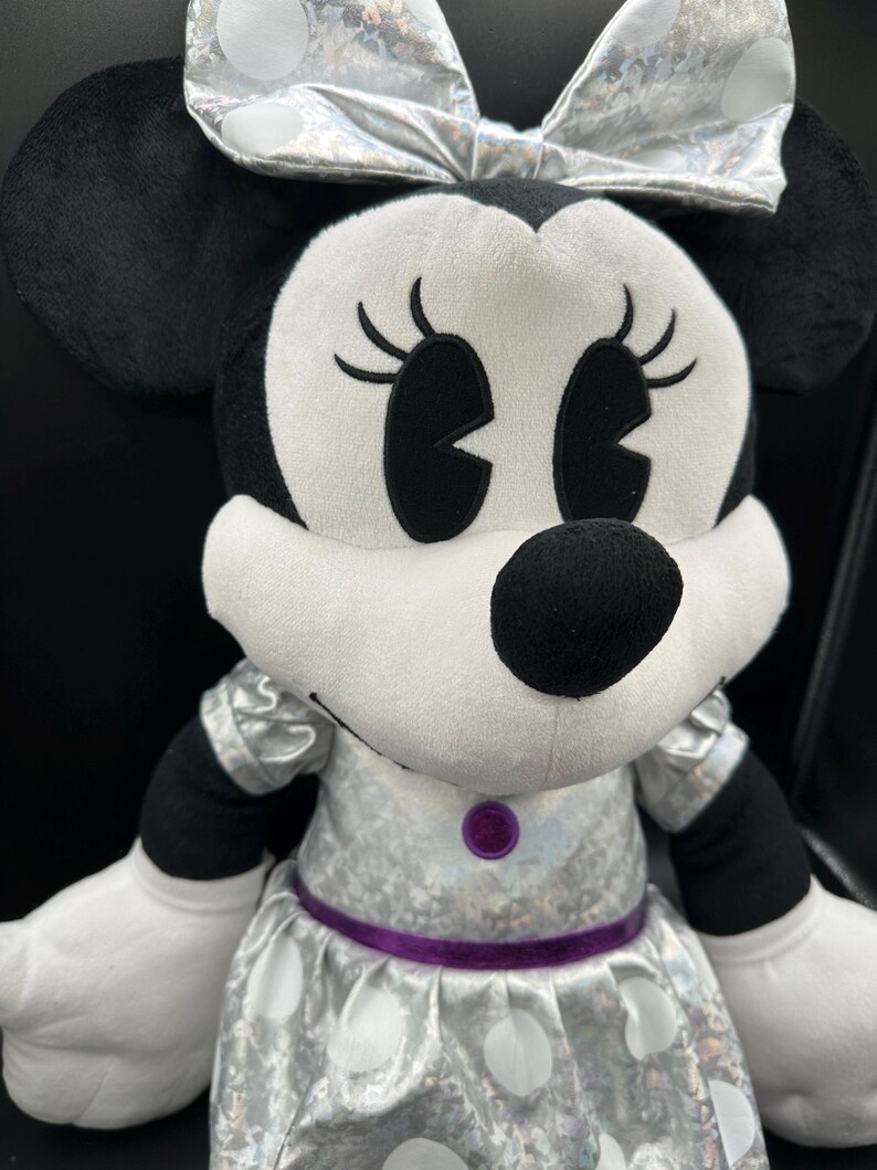 K&ouml;nnte beinhalten: Ein Pl&uuml;sch Minnie Mouse-Puppe mit silberner Schleife und Kleid mit wei&szlig;en Punkten. Die Puppe hat schwarze Ohren, Augen und Nase sowie wei&szlig;e Handschuhe. Ein lila G&uuml;rtel und Knopf akzentuieren das Kleid.