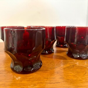 Puede incluir: Juego de seis vasos antiguos de vidrio rojo rubí. Cada vaso tiene una base texturizada única y un cuerpo cilíndrico. Los vasos están dispuestos sobre una superficie de madera, mostrando su rico color y diseño. Ideales para servir bebidas.