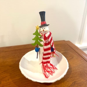 May include: A snowman-shaped candle holder with a black top hat, red and white striped scarf, and a small green Christmas tree. The snowman is holding the tree and is set in a white, scalloped-edge dish.