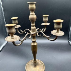 May include: A vintage brass candelabra with five candle holders. The candelabra has a central column with a round base and four curved arms extending outwards, each holding a candle cup. The brass has a warm, aged patina.