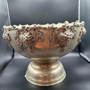 May include: A silver-toned metal pedestal bowl featuring a decorative rim adorned with embossed grape clusters and vine leaves. The bowl has a wide, circular base and a smooth, curved body that tapers toward the center, showcasing a metallic finish.