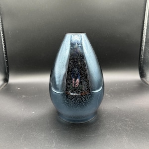 May include: A blue, teardrop-shaped vase with a speckled, glossy finish. The vase has a narrow opening and a dark interior, reflecting the surroundings. The base is a darker shade of blue.