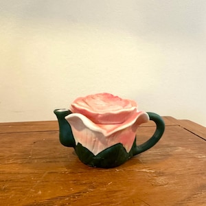 May include: A ceramic teapot shaped like a rose. The lid and flower petals are pink, the base is white, and the handle and spout are dark green. The teapot sits on a wooden surface.