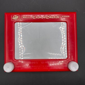 May include: A red plastic Etch A Sketch drawing toy with a rectangular frame, a grey drawing screen, and two white circular control knobs at the bottom. The frame features the text Magic Etch A Sketch Screen, Ohio Art, and The World of Toys.