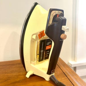 May include: A vintage General Electric iron, cream-coloured with a black handle. It has a dial for fabric settings and a "Self Clean" function. The black cord is visible. The iron is on a wooden surface.