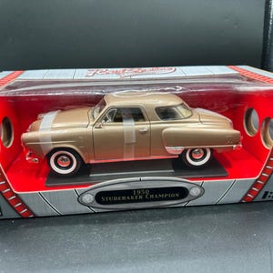 May include: A 1:18 scale die-cast model of a 1950 Studebaker Champion in a metallic gold finish. The car is secured inside its original rectangular display box with a clear plastic window, featuring the text 1950 STUDEBAKER CHAMPION on the front.