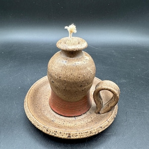 May include: A ceramic oil lamp with a speckled tan and brown finish and a reddish-brown base. The lamp features a rounded body, a narrow neck with a small opening, a central wick, and a curved handle attached to a wide, circular saucer-shaped base.