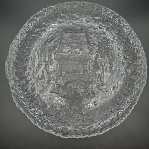 May include: A clear glass plate featuring a textured star pattern along the rim and a central embossed design depicting a fireplace scene with a hearth, a mantel, and surrounding decorative elements, all rendered in a relief style on the glass.