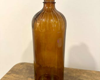 May include: A vintage amber glass bottle with a ribbed design. The bottle has a cylindrical body and a rounded shoulder, with a narrow neck and an open top. The glass has a warm, brown hue, and the bottle appears to be empty.