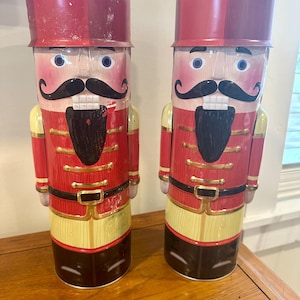 May include: Two decorative nutcracker figures. Each figure is painted with a red coat, black mustache, and a red hat. The figures have a yellow and black base with gold accents. The nutcrackers are standing upright.