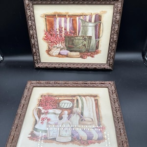 May include: Two framed wall art prints featuring bathroom scenes with pitchers, wooden buckets, scrub brushes, towels, and red berries. Each print is encased in a dark brown, ornate, textured frame with a repeating leaf pattern along the border.