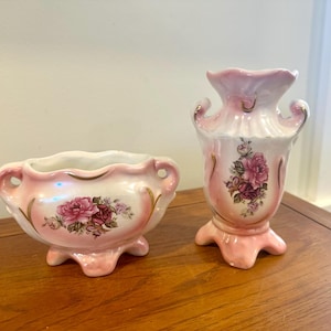 May include: A matching set of vintage ceramic vases. The pieces are light pink with gold trim and feature floral designs. The vase on the left is a small bowl with handles, and the vase on the right is a tall, decorative piece.