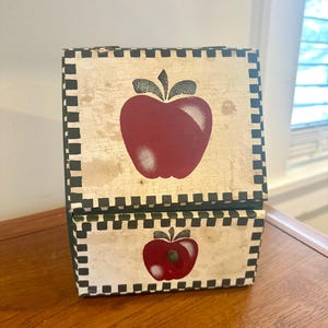 May include: A decorative wooden box with a hinged lid, featuring a hand-painted design of a red apple on a cream-colored background. The box is trimmed with a black and white checkered pattern. The box is sitting on a wooden surface.