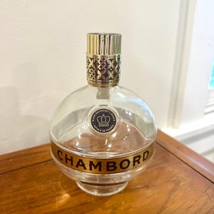 May include: A clear, round Chambord liqueur bottle with a gold-colored cap and label. The label reads "CHAMBORD" in gold lettering. The bottle has a distinctive shape and is made of glass.