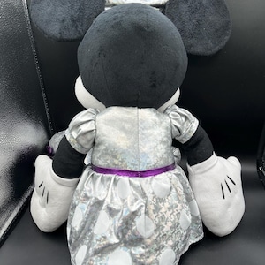 K&ouml;nnte beinhalten: Eine Pl&uuml;schpuppe von Minnie Mouse, von hinten gesehen, mit schwarzen Ohren und Kopf, einem silbernen Kleid mit wei&szlig;en Punkten und einer passenden Schleife. Die Puppe hat wei&szlig;e Handschuhe und einen lila G&uuml;rtel.