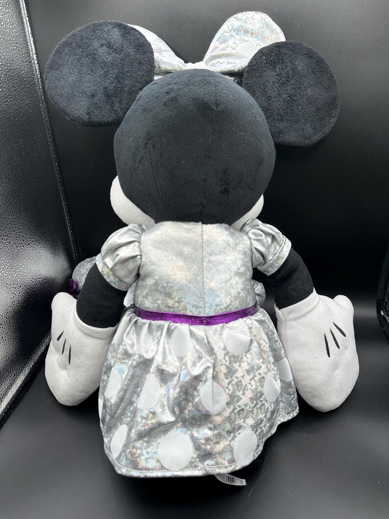 K&ouml;nnte beinhalten: Eine Pl&uuml;schpuppe von Minnie Mouse, von hinten gesehen, mit schwarzen Ohren und Kopf, einem silbernen Kleid mit wei&szlig;en Punkten und einer passenden Schleife. Die Puppe hat wei&szlig;e Handschuhe und einen lila G&uuml;rtel.
