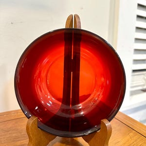 May include: A striking red glass plate, presented on a wooden stand. The plate's colour is a deep, rich red, with a simple, elegant design. The wooden stand offers a warm contrast to the glass. A decorative item for the home.