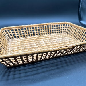 May include: A rectangular, light brown woven basket with an open grid pattern. The basket features a flat base and slightly angled sides, constructed from thin, flexible material strips arranged in a consistent, repeating crisscross weave.