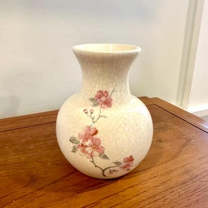 Puede incluir: Jarrón de cerámica color crema con un esmalte craquelado y un diseño floral. El jarrón tiene un cuerpo redondeado y un cuello acampanado. Flores rosas y hojas verdes adornan la superficie, añadiendo elegancia a la pieza decorativa.