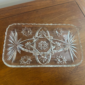 May include: A clear, rectangular glass dish with a starburst and star pattern. The edges of the dish are scalloped. The dish is on a wooden surface.