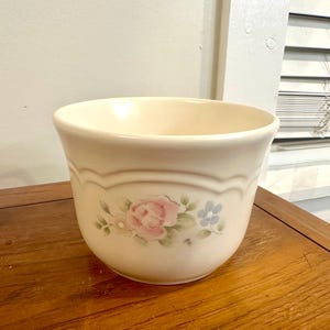 May include: Cream-colored ceramic bowl with a floral design. The bowl features a pink rose and blue flowers with green leaves. The rim has a decorative brown border. The bowl is sitting on a wooden surface.