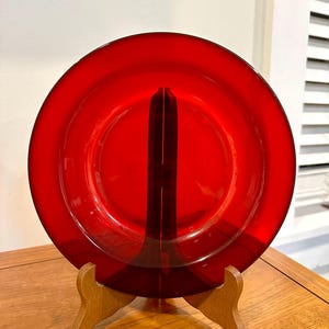 May include: A vibrant red glass plate displayed on a wooden stand. The plate is round with a slightly raised edge. The wooden stand has a simple, classic design. The plate is the focal point, showcasing its rich color and reflective surface.