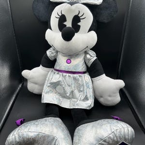 K&ouml;nnte beinhalten: Eine Pl&uuml;schpuppe von Minnie Mouse mit einem silbernen Kleid und einer Schleife. Die Puppe hat schwarze Ohren, eine schwarze Nase und wei&szlig;e Handschuhe. Das Kleid hat wei&szlig;e Punkte und eine lila Taille. Die Schuhe der Puppe sind silberfarben und haben eine lila Schleife.