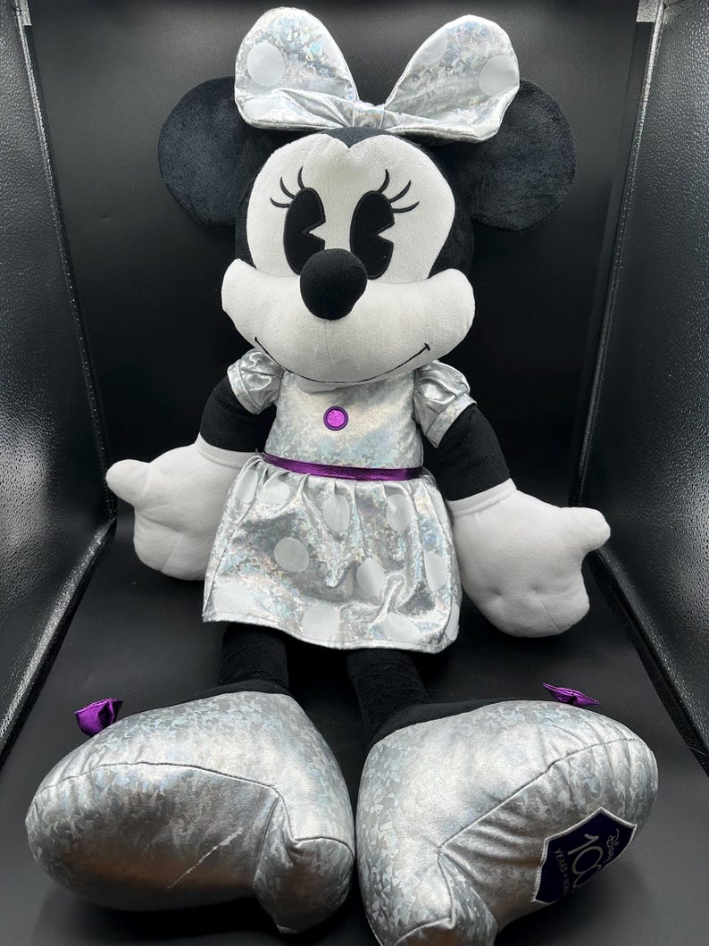 K&ouml;nnte beinhalten: Eine Pl&uuml;schpuppe von Minnie Mouse mit einem silbernen Kleid und einer Schleife. Die Puppe hat schwarze Ohren, eine schwarze Nase und wei&szlig;e Handschuhe. Das Kleid hat wei&szlig;e Punkte und eine lila Taille. Die Schuhe der Puppe sind silberfarben und haben eine lila Schleife.