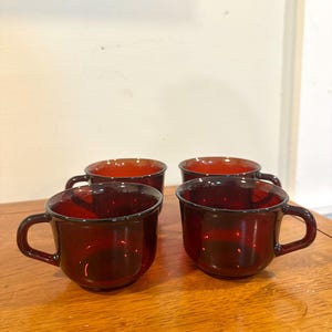 May include: Four ruby red glass coffee cups with handles are arranged on a wooden surface. The cups have a simple, rounded design and a deep red color, creating a vintage aesthetic. The cups are empty.