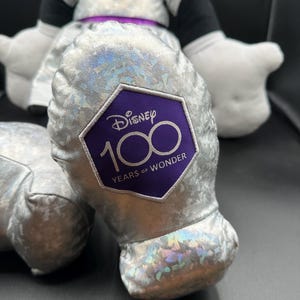 K&ouml;nnte beinhalten: Eine Pl&uuml;schpuppe von Minnie Mouse in einem silbernen Kleid mit lila Akzenten. Der Fu&szlig; der Puppe hat einen lila Aufn&auml;her mit dem Disney-Logo und dem Text "100 Years of Wonder."