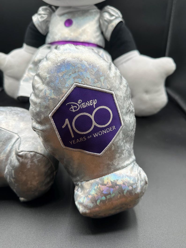 K&ouml;nnte beinhalten: Eine Pl&uuml;schpuppe von Minnie Mouse in einem silbernen Kleid mit lila Akzenten. Der Fu&szlig; der Puppe hat einen lila Aufn&auml;her mit dem Disney-Logo und dem Text "100 Years of Wonder."