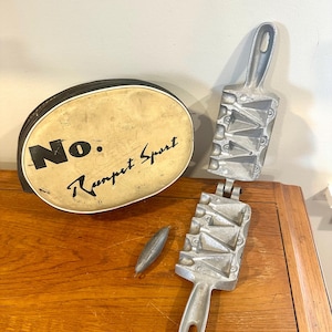 May include: An oval, cream-colored case with the text "No. Rampet Sport" sits next to a metal fishing weight and a two-part metal fishing weight mold. The mold is open, revealing the cavities for the weights.