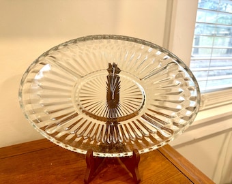 May include: A clear, oval-shaped glass serving dish with three compartments, resting on a dark wooden stand. The dish features a ribbed design radiating from the center. The stand is made of dark wood.