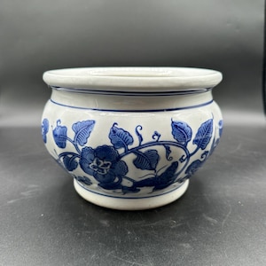 May include: A white ceramic bowl with a rounded body and a flared rim, featuring a hand-painted cobalt blue floral and vine pattern that wraps around the exterior. The base is slightly tapered, and the design includes detailed leaves and blossoms.