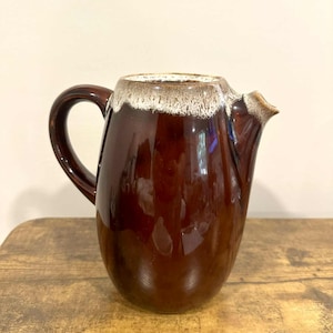 May include: A brown ceramic pitcher with a handle and spout. The pitcher has a glossy finish and a lighter-colored rim. The pitcher is sitting on a wooden surface.