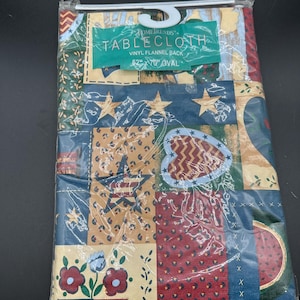 May include: A vinyl flannel back oval tablecloth measuring 52 inches by 70 inches, packaged in clear plastic. The fabric features a multi-colored patchwork design with stars, hearts, flowers, and geometric patterns in blue, red, and tan.