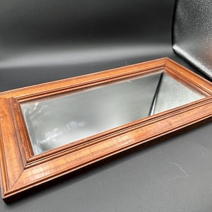 May include: A rectangular mirror featuring a medium-toned brown wooden frame with a multi-layered, stepped profile. The glass surface is clear and reflective, set within the structured, angled edges of the wood border, creating a clean, linear design.