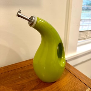 May include: A vibrant green ceramic oil dispenser with a stainless steel spout and a small handle. The dispenser has a curved design with a circular opening on the side. It sits on a wooden surface.