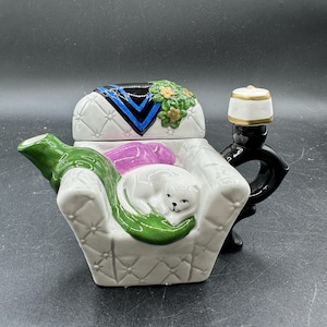 May include: A ceramic teapot shaped like a white tufted armchair with a green spout, a black handle topped with a white and gold fez, and a lid featuring a blue and black patterned throw blanket, small yellow flowers, and a sleeping white cat.