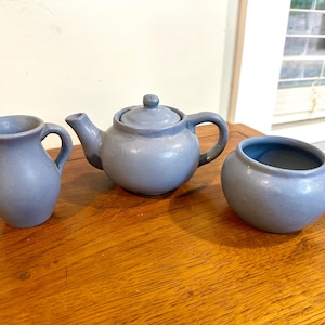 May include: A light blue ceramic tea set, including a teapot with a lid, a creamer pitcher, and a sugar bowl. The set is displayed on a wooden surface. The teapot has a handle and spout. The sugar bowl is round.